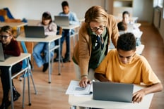 Student at desk and working on his Chromebook in a classroom with a teacher looking over his shoulder